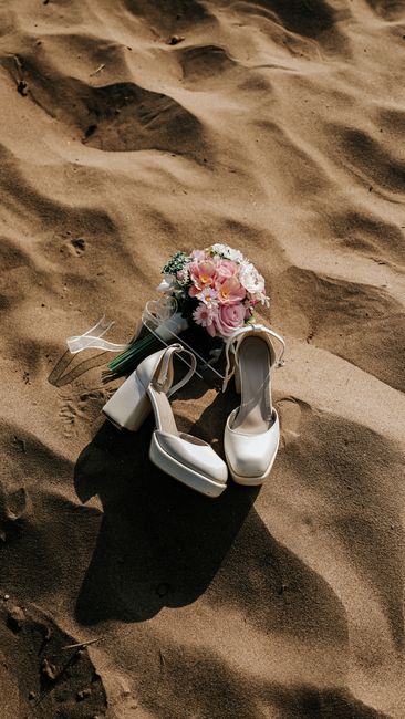 A pair of white satin wedding shoes with rounded toes and chunky heels rest on the golden sandy beach, casting soft shadows. Beside the shoes, a delicate bridal bouquet featuring pink roses, white hyd