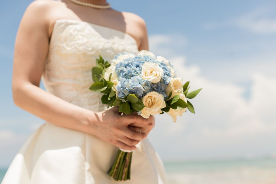 A bride holding a bouquet arrangement of light blue hydrangeas and cream roses, accented with lush green leaves, wrapped with a simple green ribbon. She is wearing a strapless ivory wedding dress with
