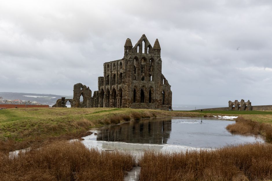 An overcast sky overlooks the ruins of Whitby Abbey in North Yorkshire, with weathered stone walls and arches standing amidst grassy terrain. A small, still body of water in the foreground reflects pa