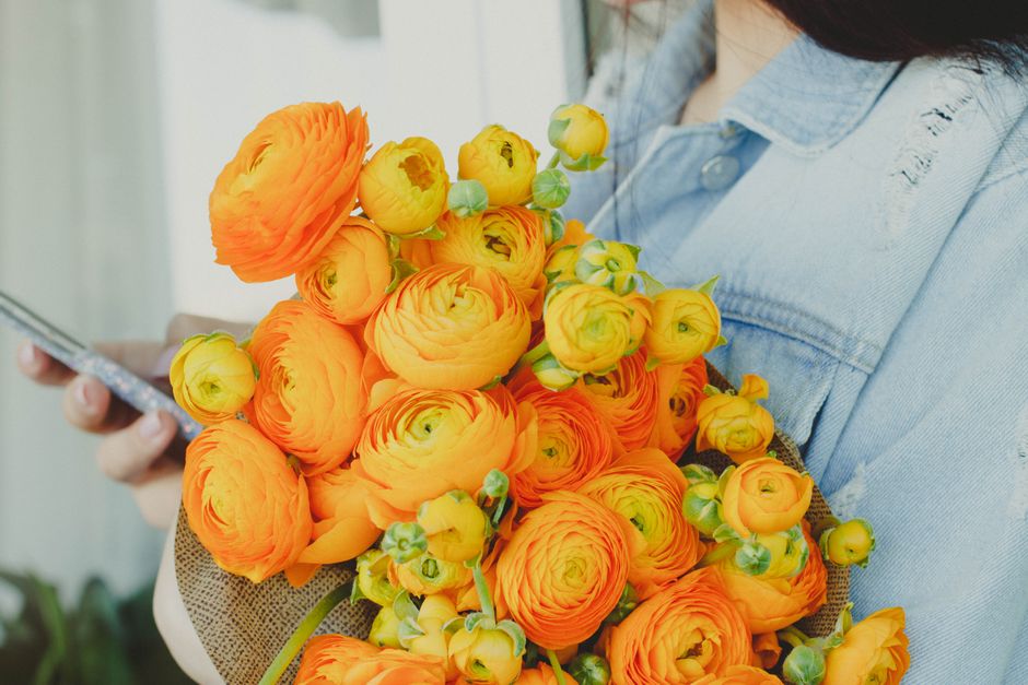A person wearing a light blue denim shirt holds a large bouquet of vibrant orange and yellow ranunculus flowers with lush green buds and stems. The flowers are arranged in a dense, rounded cluster, sh